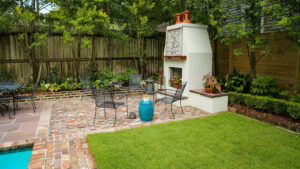 Brick patio with an outdoor fireplace beside a lawn and layered garden beds in Uptown New Orleans