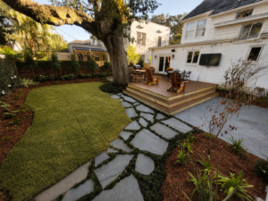 Audubon Boulevard rear garden with a mature live oak, new lawn, layered planting, flagstone path, and wooden deck.