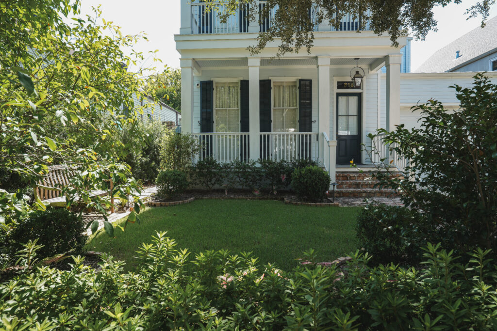 Maintained front garden with a central lawn, layered evergreen planting, brick edging, and a shaded bench at a traditional New Orleans residence.