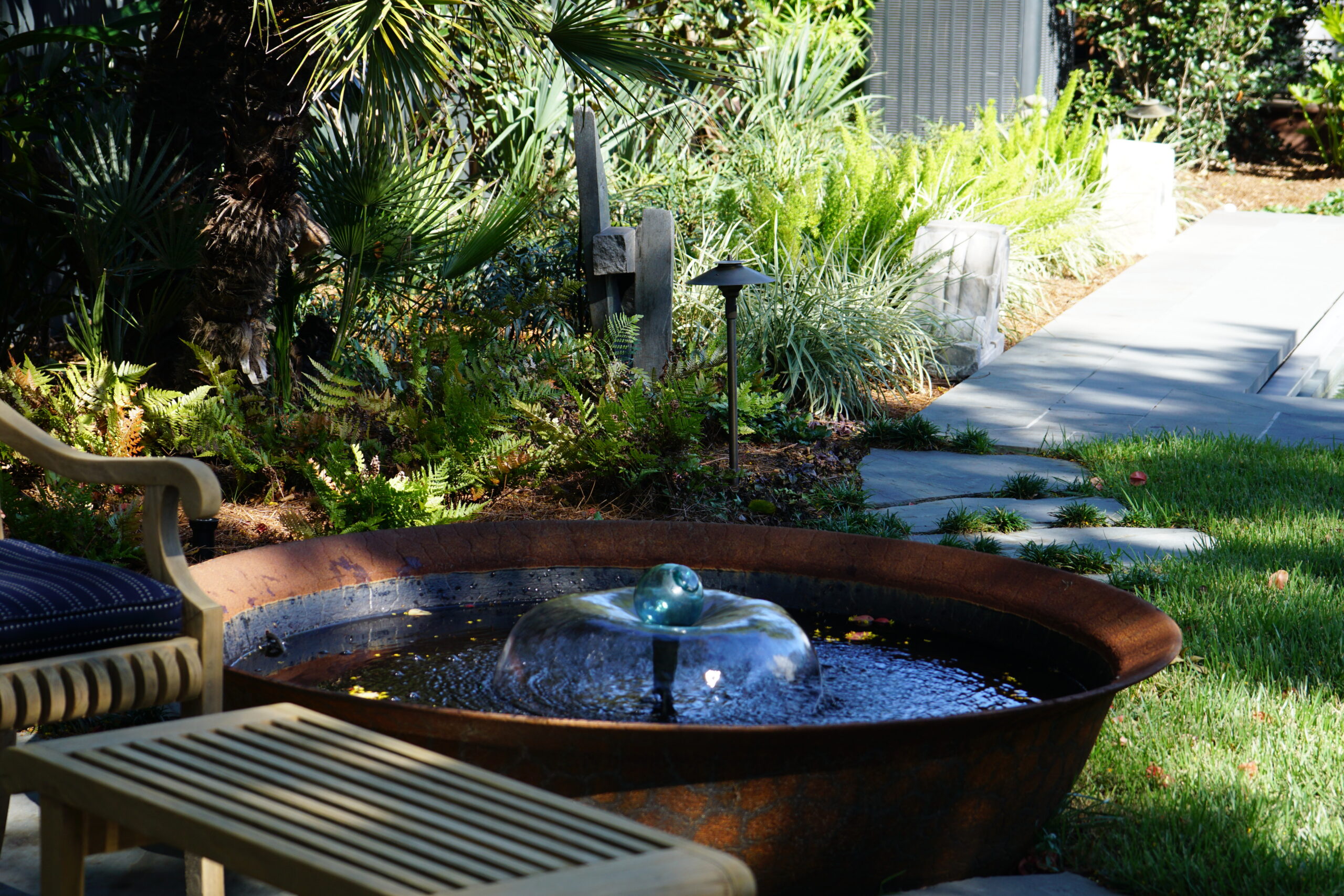 Round bubbling water feature surrounded by palms and tropical planting in an Uptown New Orleans garden