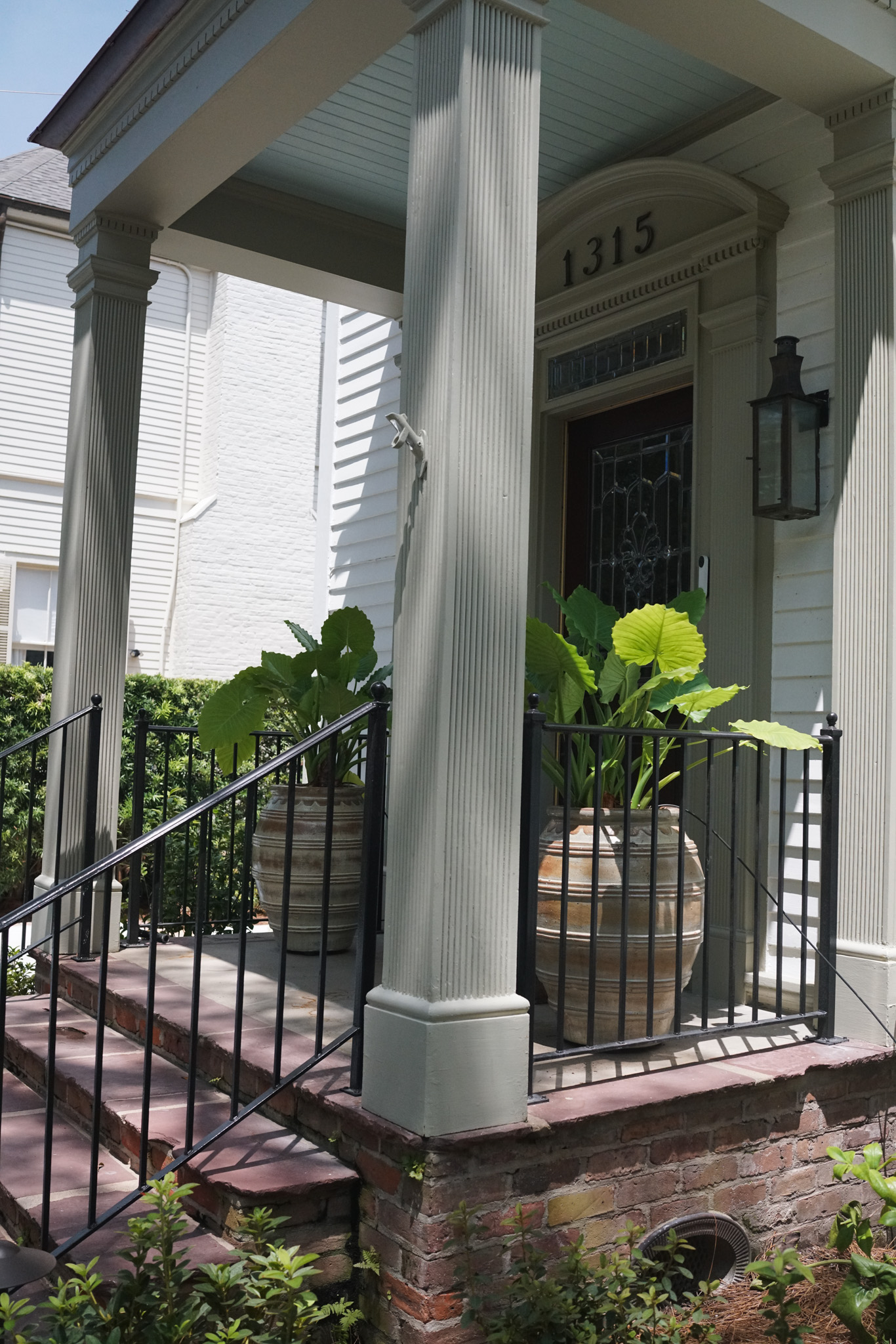 Traditional State Street front porch entrance with brick steps, classical columns, iron railings, and large planters filled with tropical foliage.