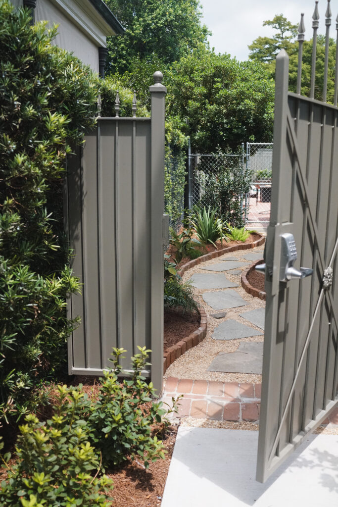Open custom garden gate leading to a curved flagstone path with brick edging and layered plantings in an Uptown New Orleans secret garden