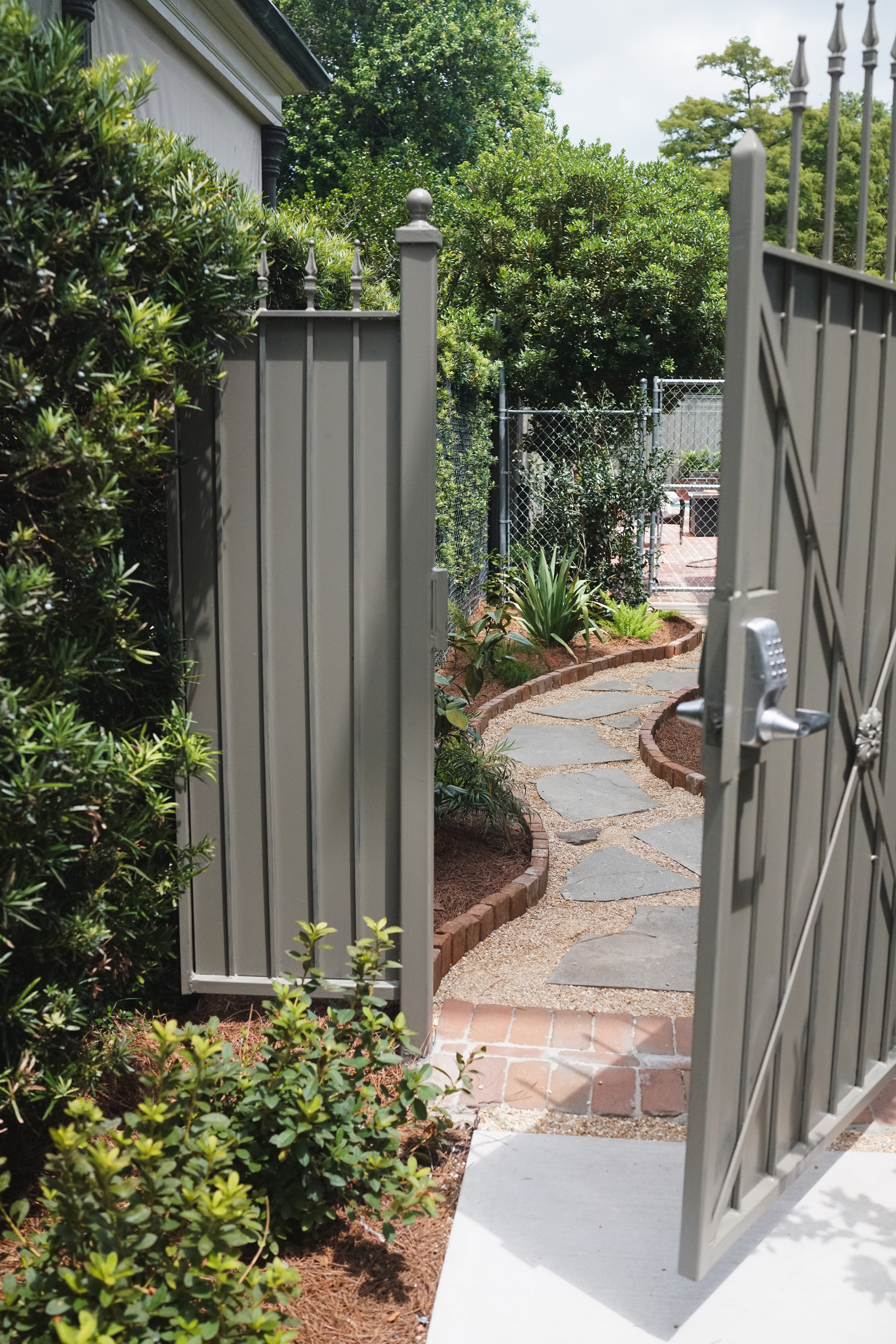 Open custom garden gate leading to a curved flagstone path with brick edging and layered plantings in an Uptown New Orleans secret garden