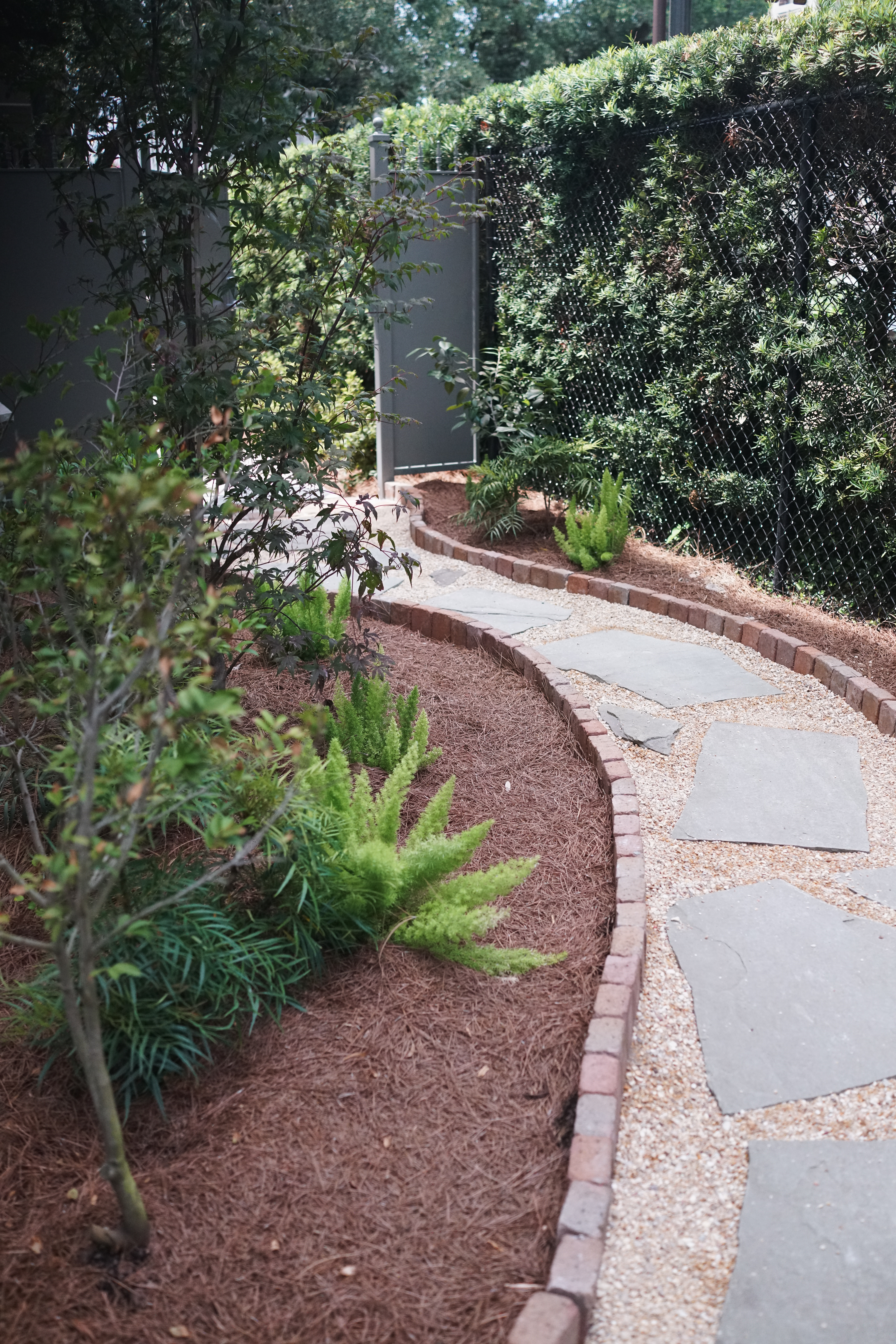 Curved secret garden path with irregular flagstone, crushed stone, brick edging, Japanese maple, ferns, and layered evergreen plantings in Uptown New Orleans.