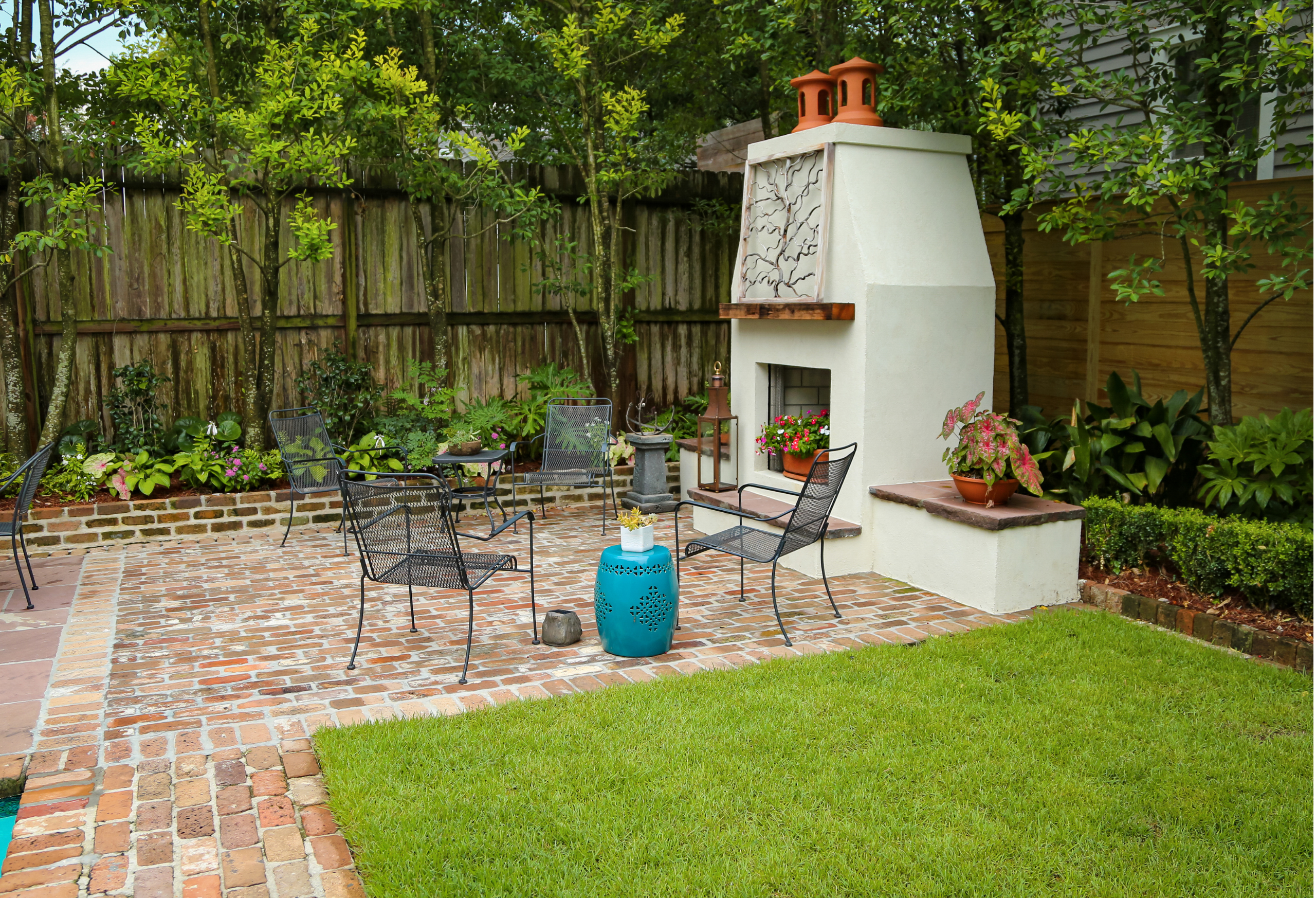 Brick patio with black metal seating and a white outdoor fireplace surrounded by lush garden plantings in Uptown New Orleans