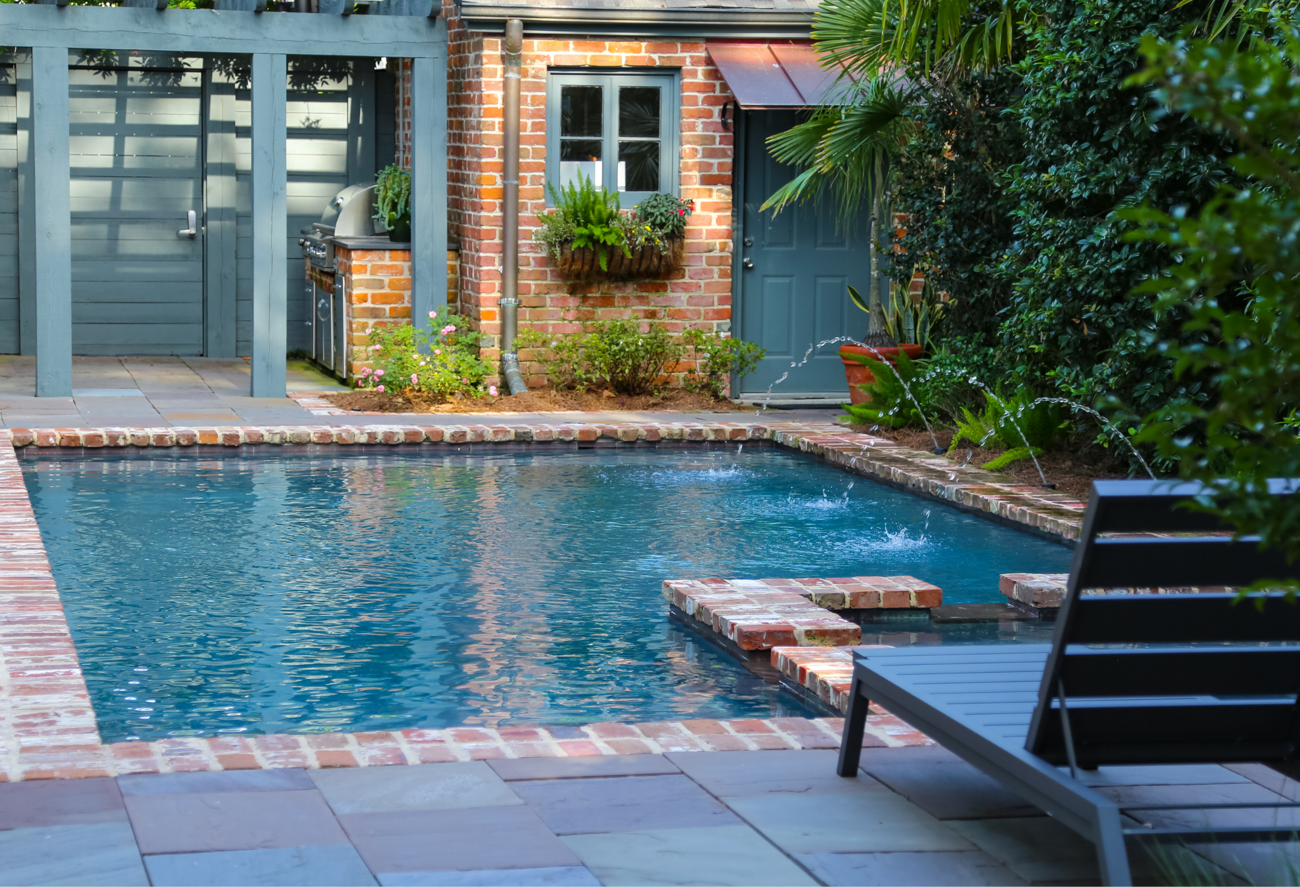 Brick-edged swimming pool with water features, lounge seating, and a shaded outdoor kitchen in a Metairie backyard