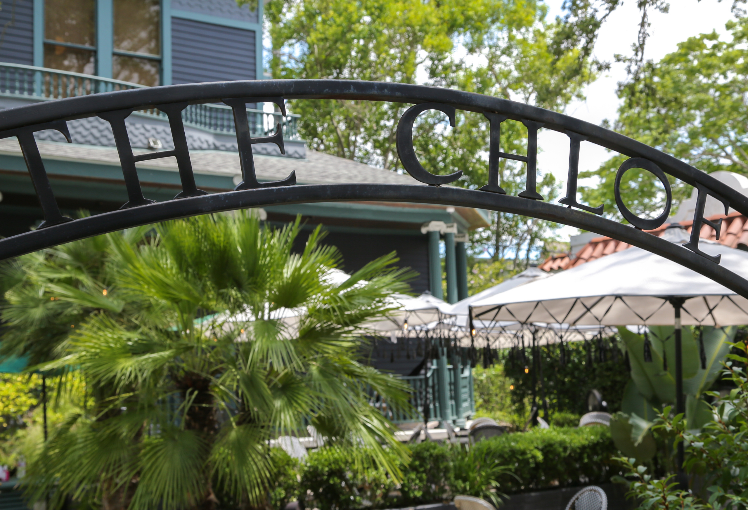 Decorative iron arch at The Chloe courtyard entrance, framing palms, patio umbrellas, and lush garden plantings in New Orleans