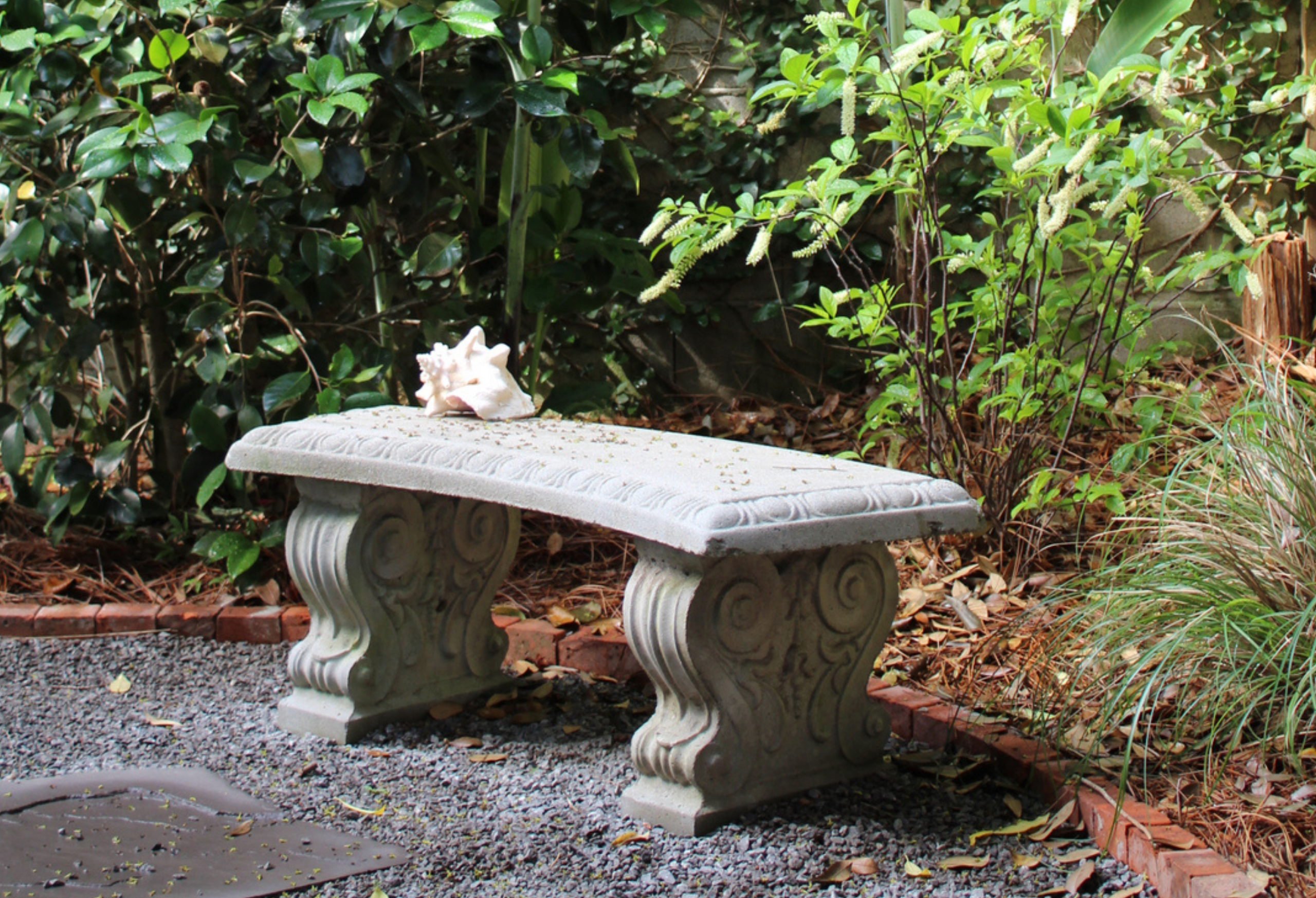 Ornate stone garden bench on a gravel path surrounded by lush plantings and brick edging