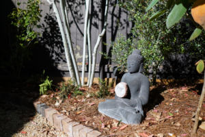 Dark Buddha garden statue holding a stone beside brick edging, mulch, and layered planting in the Constance Street garden.