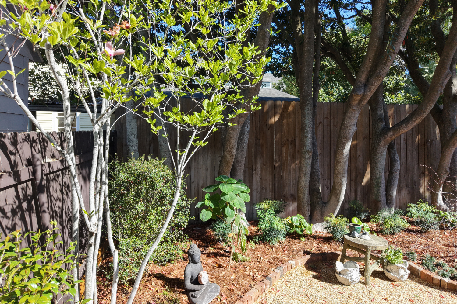 Dark Buddha garden statue holding a stone beside brick edging, mulch, and layered planting in the Constance Street garden.