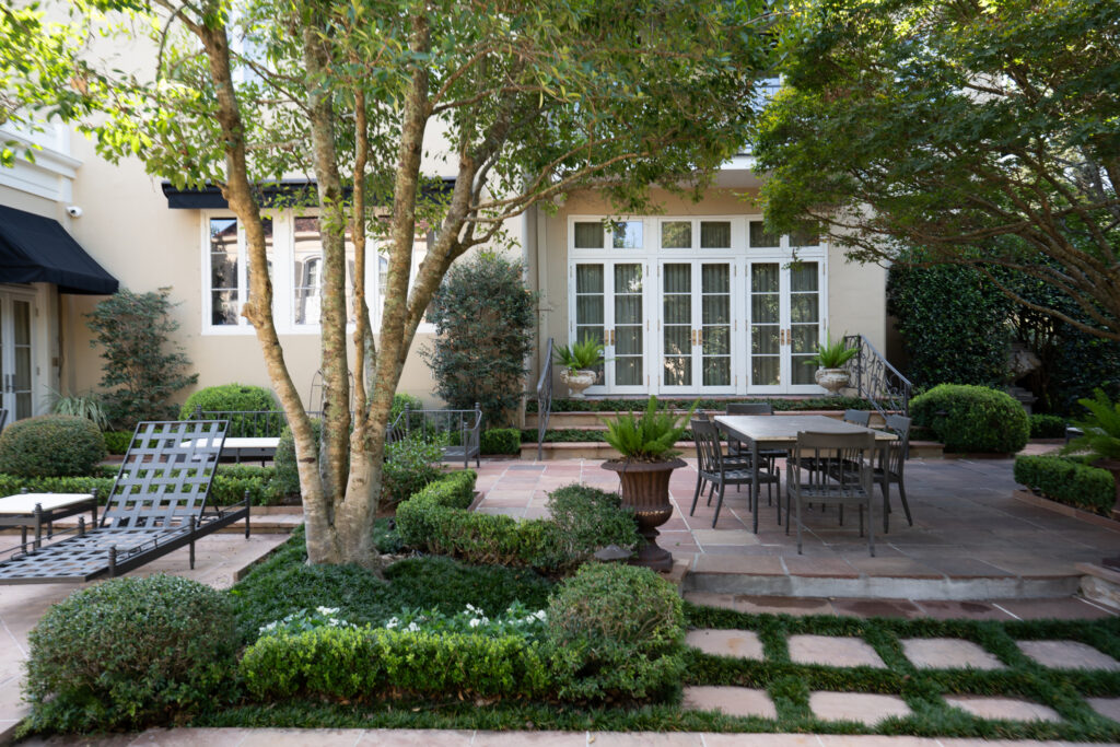 New Orleans residential courtyard with a stone dining terrace, clipped planting, mature trees, and French doors opening from the home.