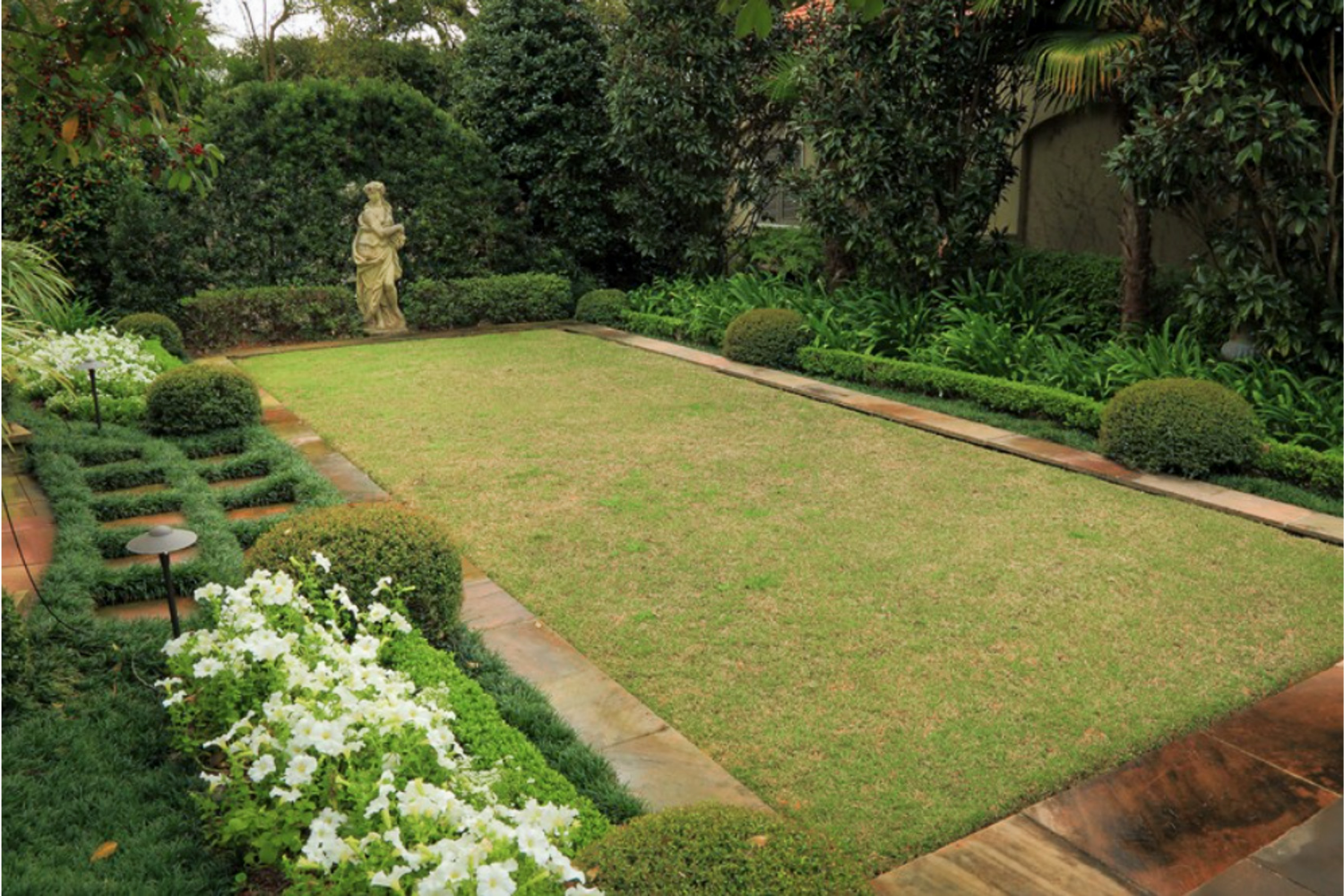 Formal landscape design with classical statue, white flowers, clipped shrubs, lawn, and layered evergreen planting.