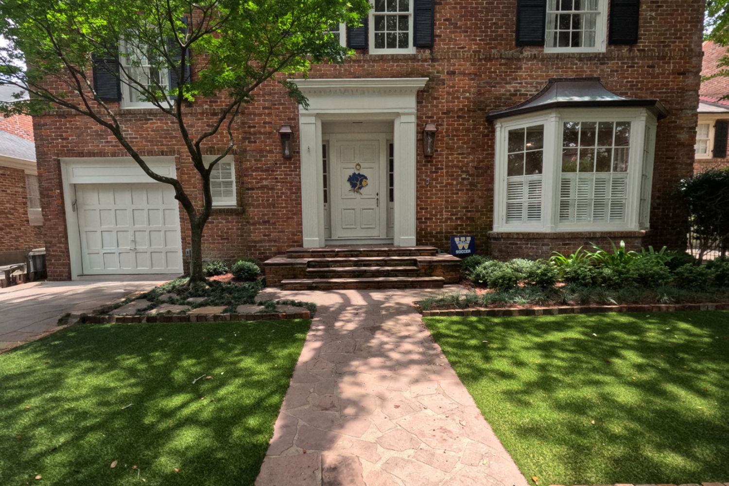 Front entrance of a brick Uptown New Orleans home with a flagstone walk, lawn panels, brick-edged planting beds, and mature trees at Nashville Avenue Garden.