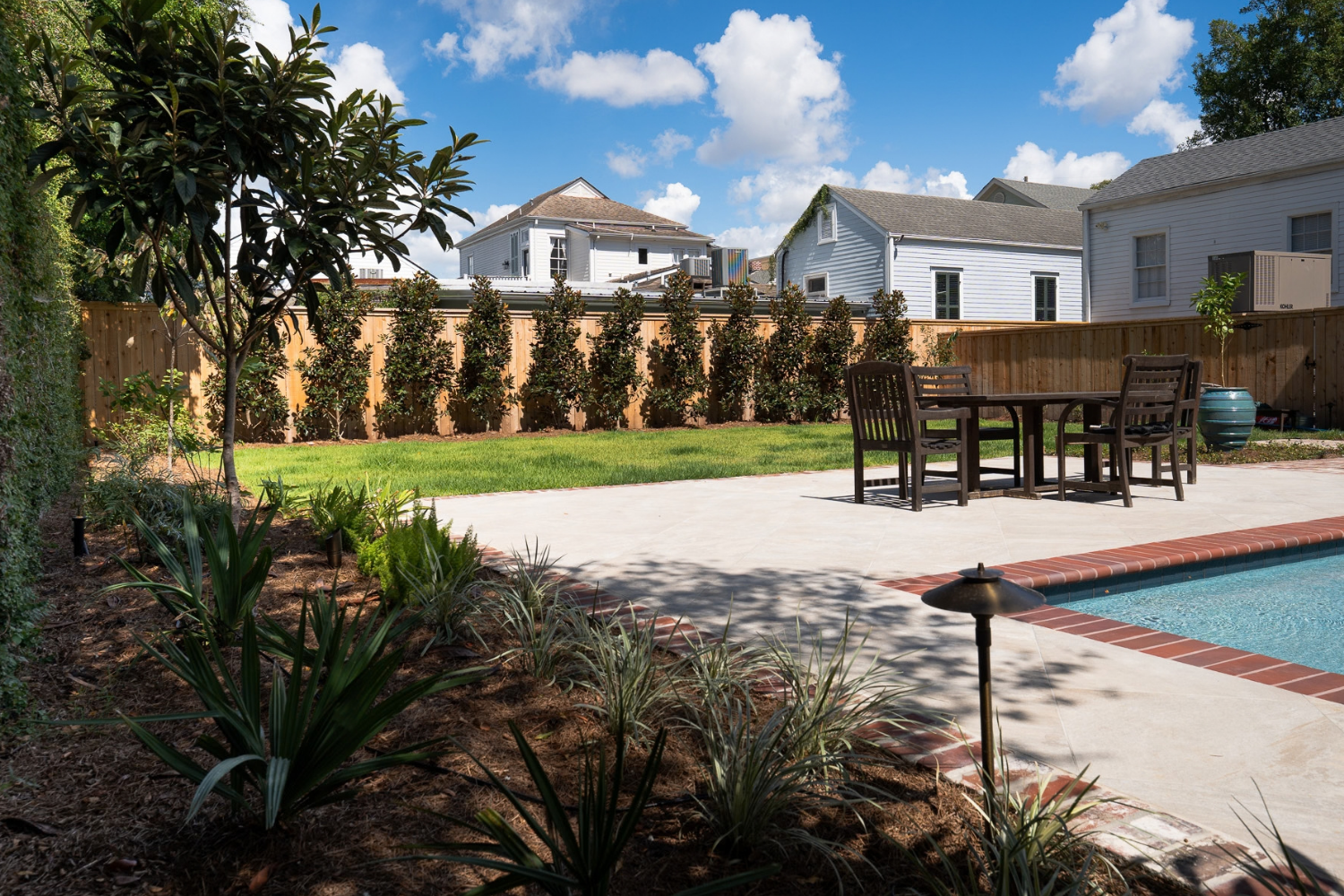 New Orleans pool garden with brick coping, planting beds, a wood privacy fence, and outdoor dining area.